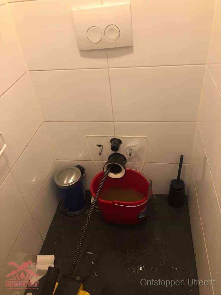 Een ontstopte wand-wc met verwijderde toiletpot waar een ontstoppingsveer in de wanduitgang verdwijnt, het achterhalen waar de verstopping zit in Utrecht