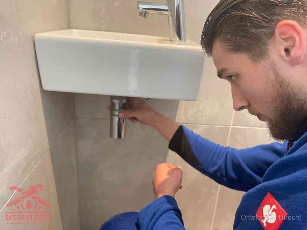 Een monteur draait met de hand de zwanenhalssifon onder een fonteinwasbak in de toiletruimte los om de bron van rioollucht in Utrecht te vinden