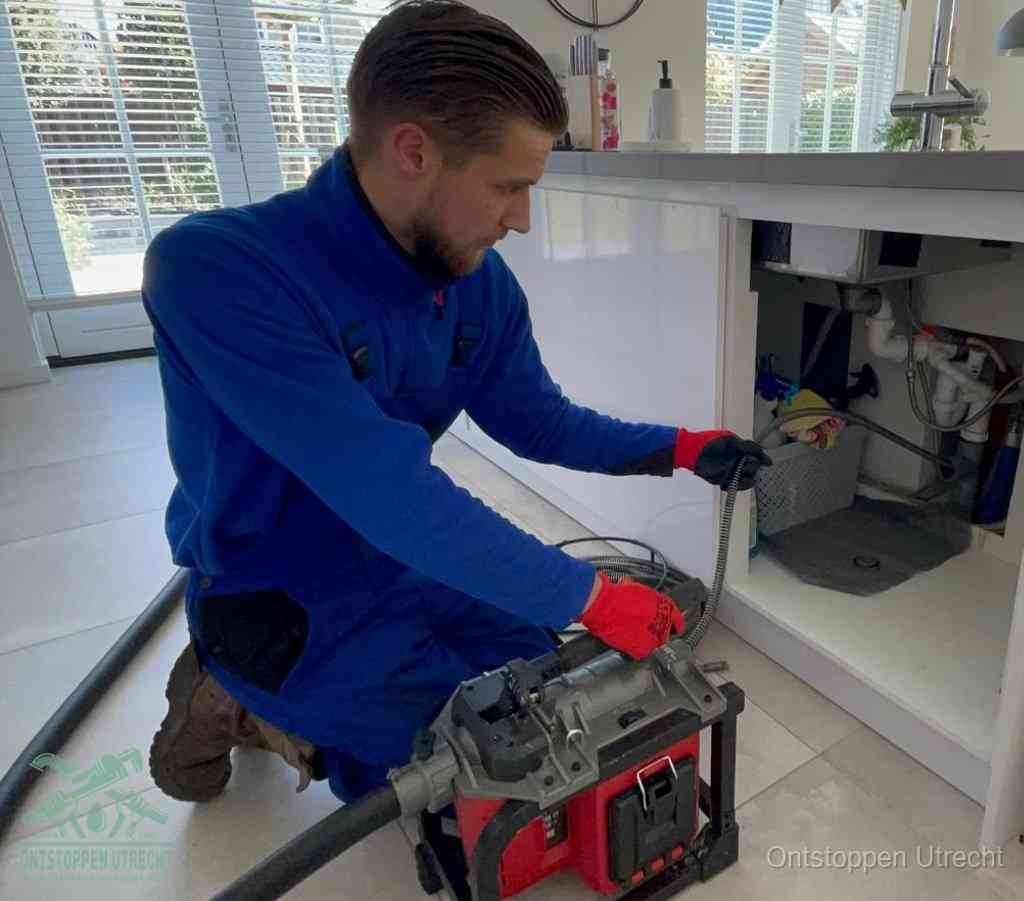 Een monteur met rode handschoenen knielt in een keuken en voert een elektrische ontstoppingsveer in tijdens het professioneel afvoer ontstoppen in Utrecht